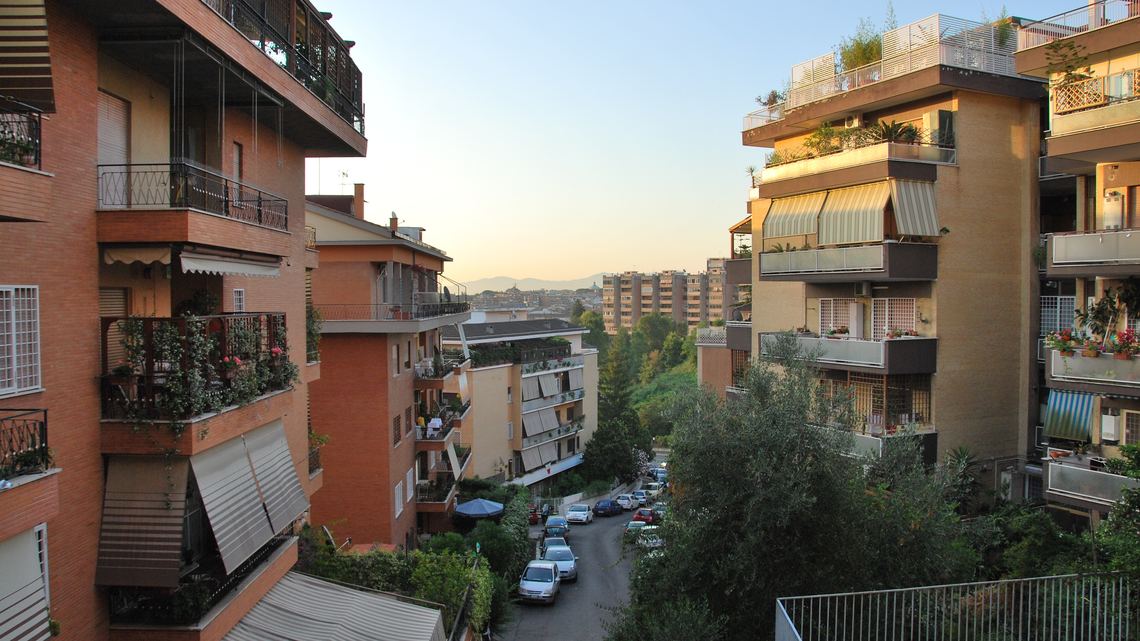 Via Enrico Besta al tramonto, vista dall'alto: palazzine di mattoni e intonaco con i balconi pieni di piante e le tende da sole, auto in sosta lungo la strada che scende, alberi, e in fondo i tetti di Roma e le colline