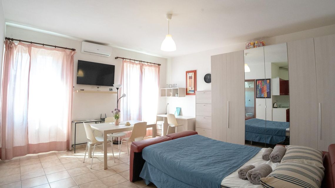 Il monolocale di Furio Camillo: letto con il copriletto blu, tavolo con le sedie, armadio a specchio e due finestre