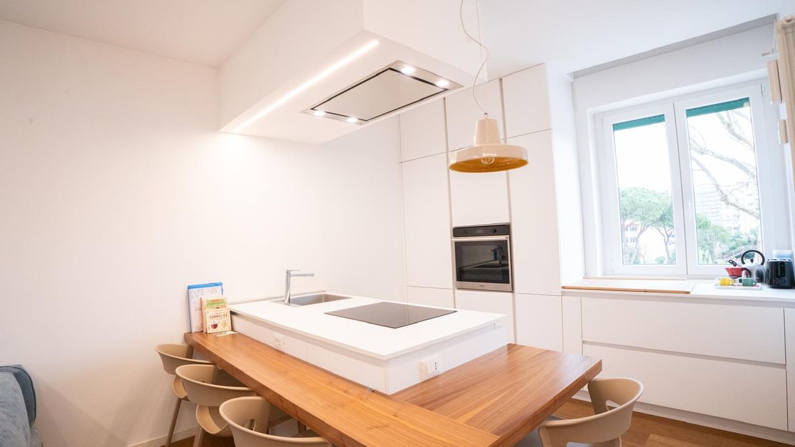 La cucina dell'appartamento al Tuscolano: isola bianca, tavolo lungo di legno, lampada gialla e la finestra sui pini